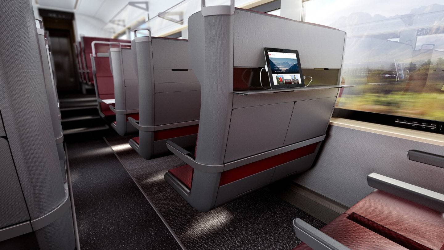 Train seats viewed from a seated position across the aisle. On the back of two seats, you can see a shelf, with a tablet leaning on it