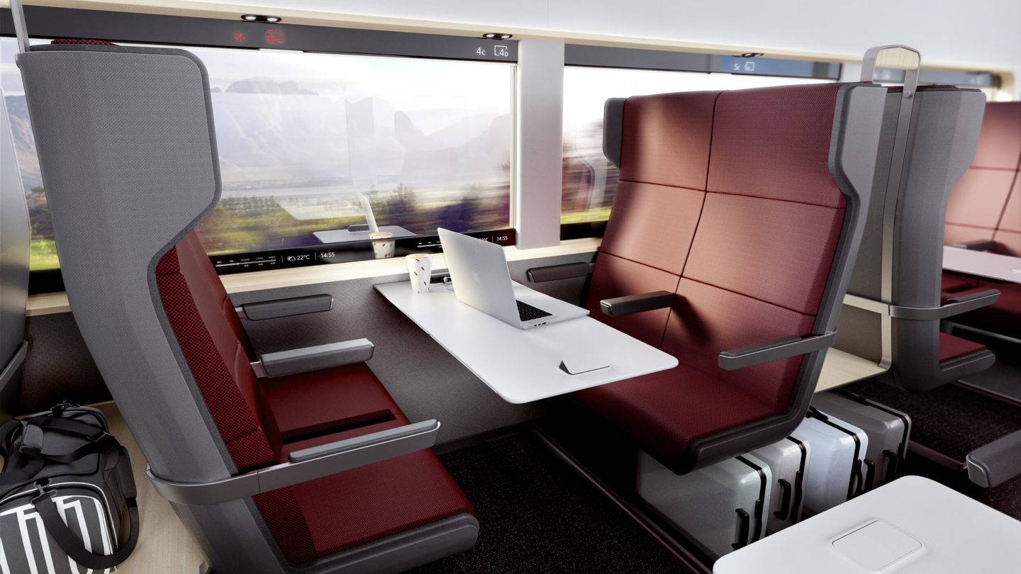 Four train seats facing one another. A large table features in the middle, with a laptop placed on it. Between this group of four seats and the adjacent ones is a storage space, where you can see bags and carry on luggage neatly stowed