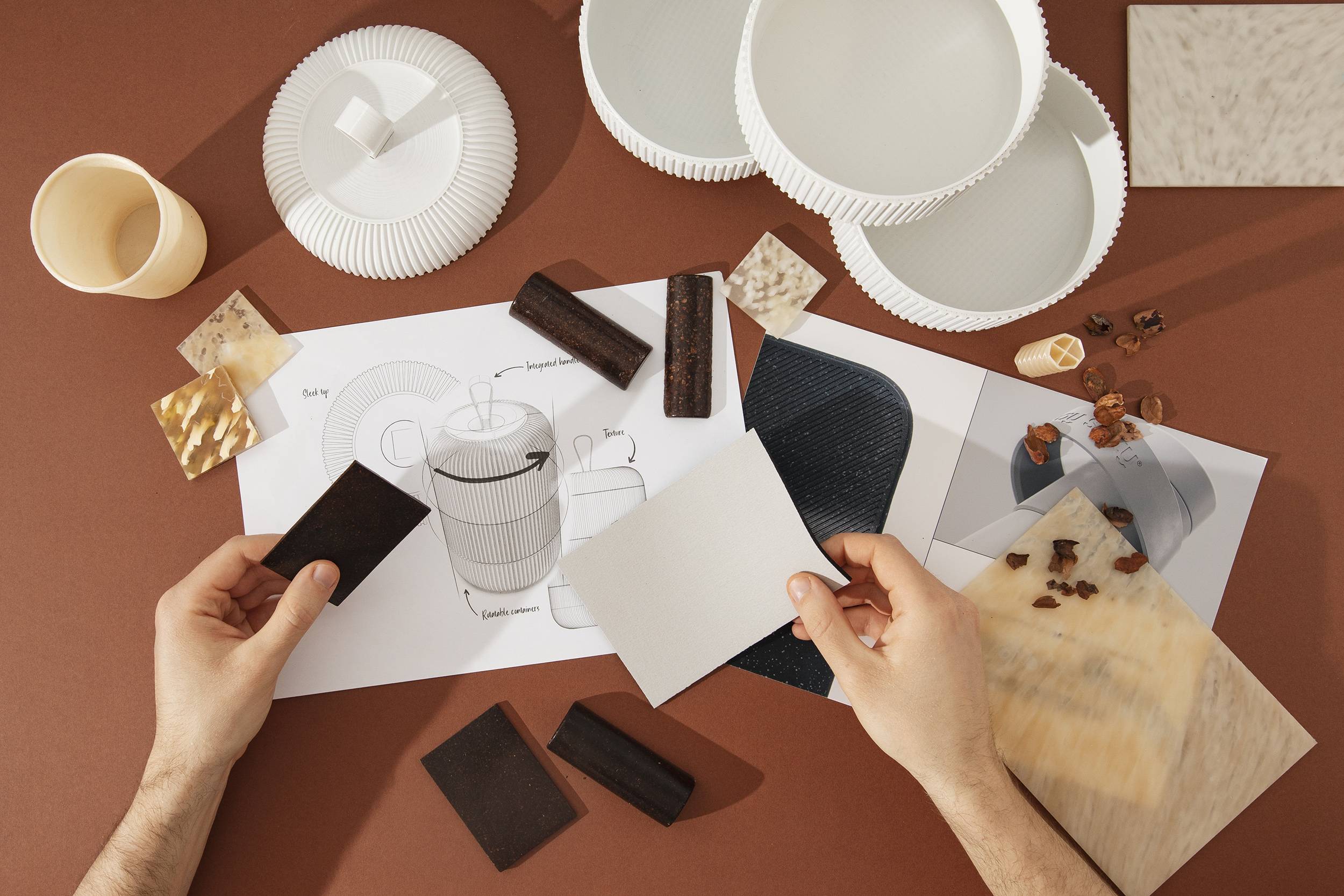 Two hands shown holding material samples over a series of sketches, drawings, material samples and 3D models